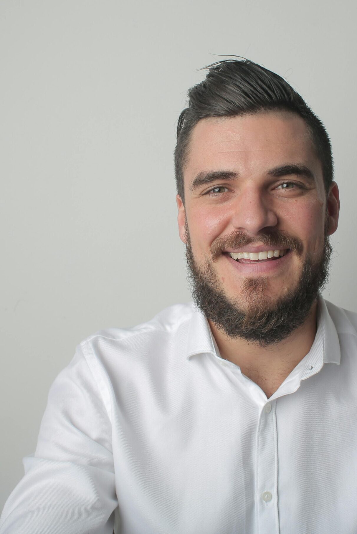 Portrait of a young bearded man smiling, wearing a white shirt indoors against a gray background.