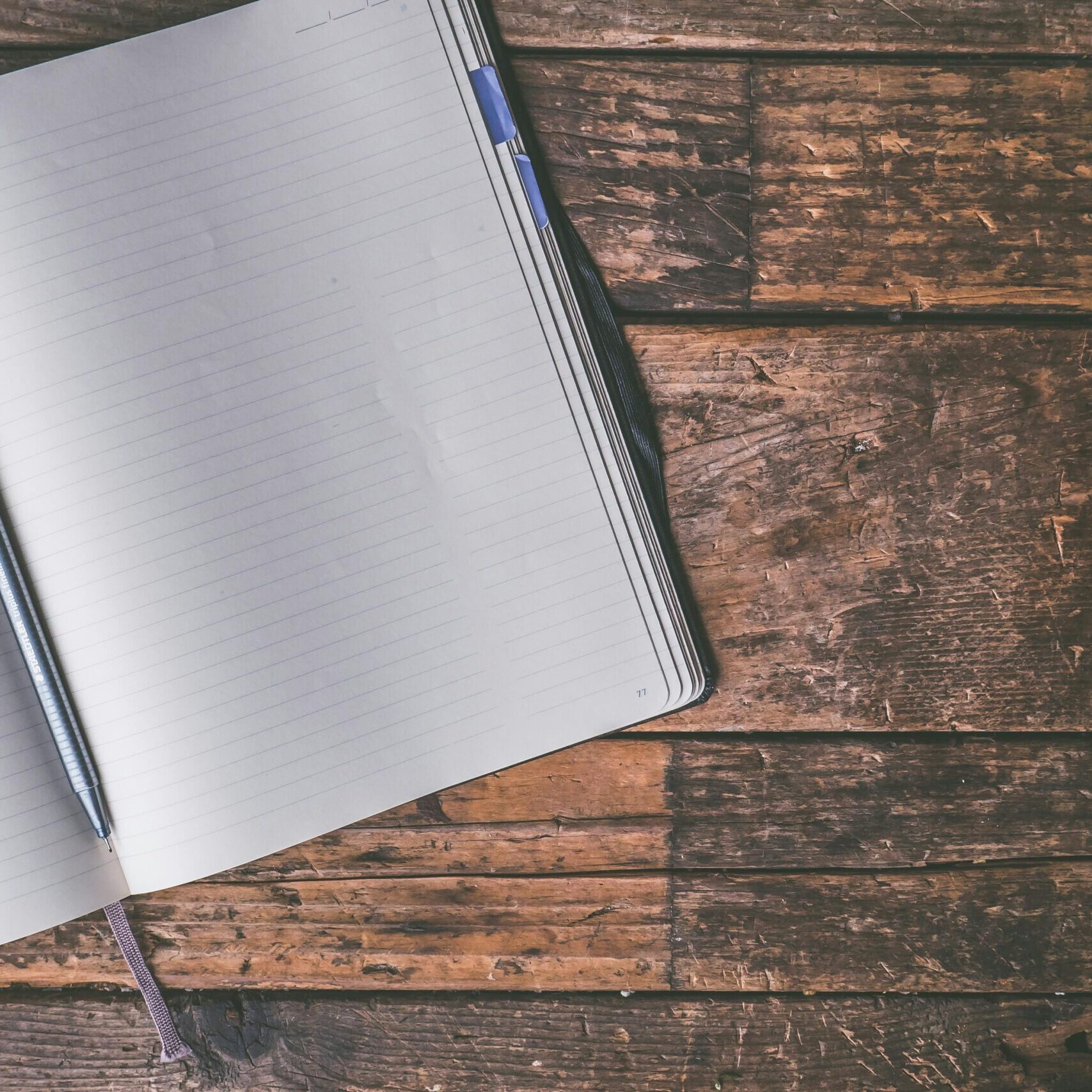 An open notebook with a blue pen lies on a rustic wooden table, inviting creativity and writing.